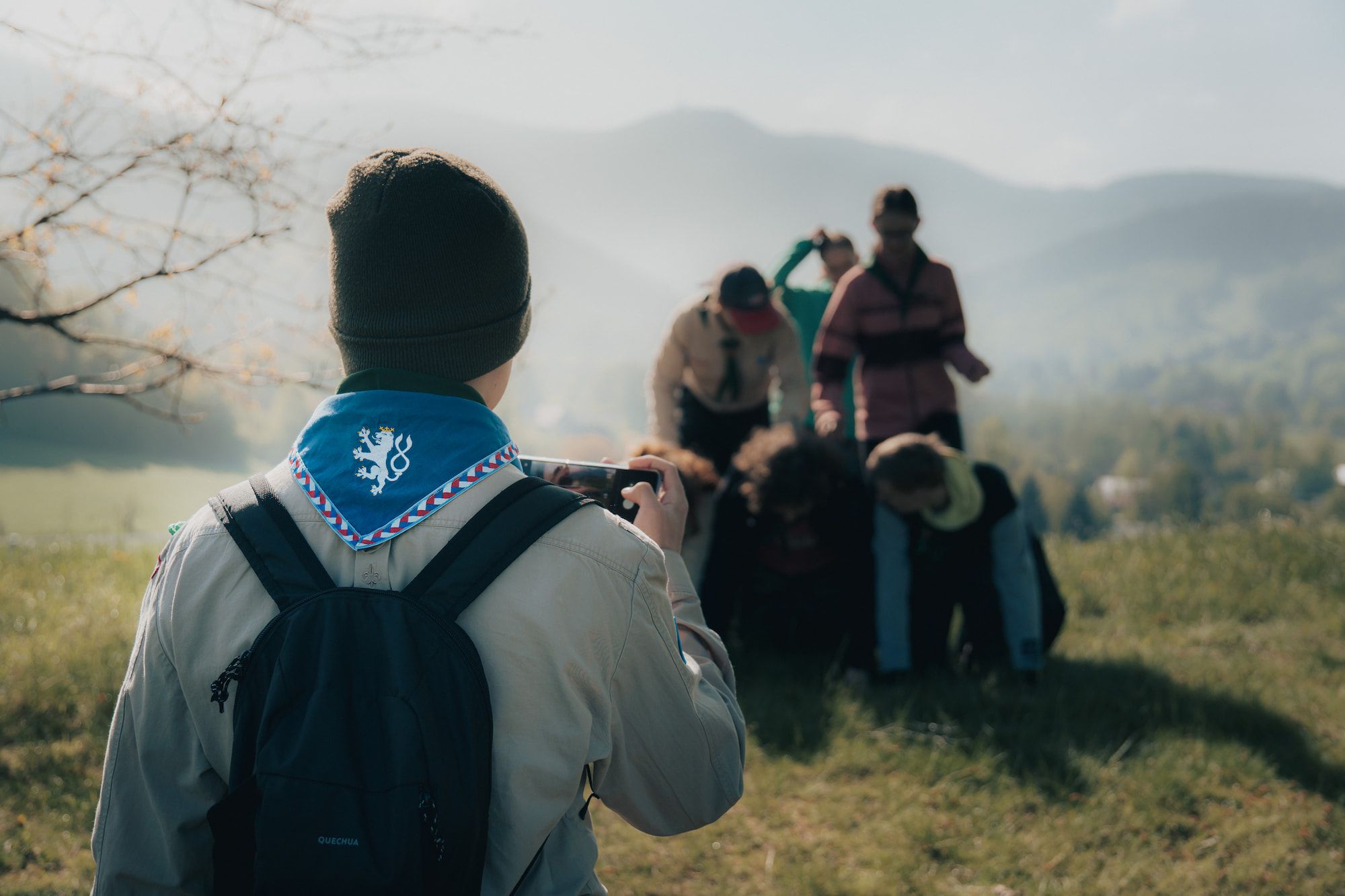 Osoba s batohem a šátkem fotografuje skautské děti, které tvoří lidskou pyramidu na travnatém kopci, v pozadí jsou hory.