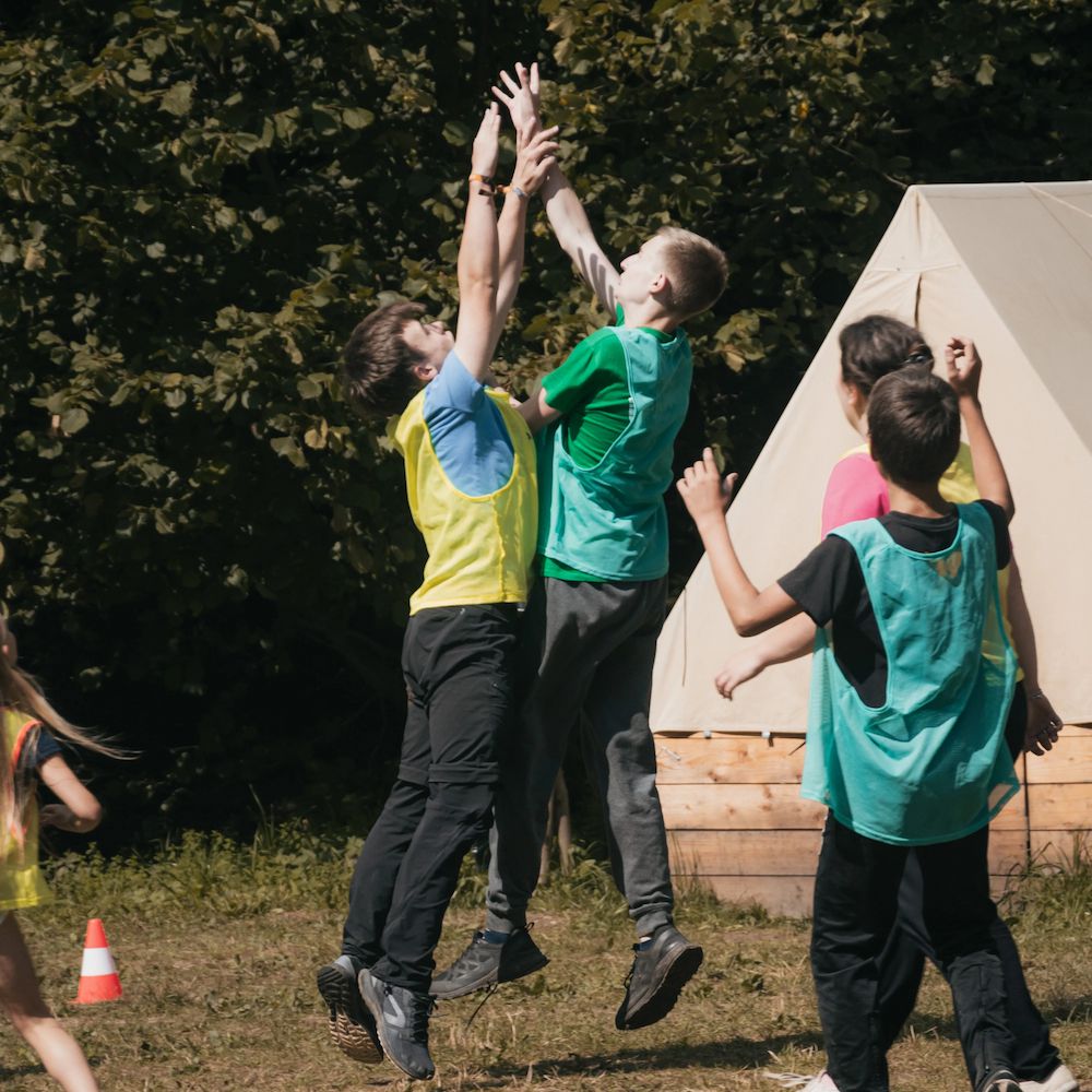 Děti energicky skáčou, aby chytily míč na travnatém hřišti obklopeném stromy a stany. Mají na sobě pestrobarevné sportovní vesty, které vytvářejí hravou atmosféru.