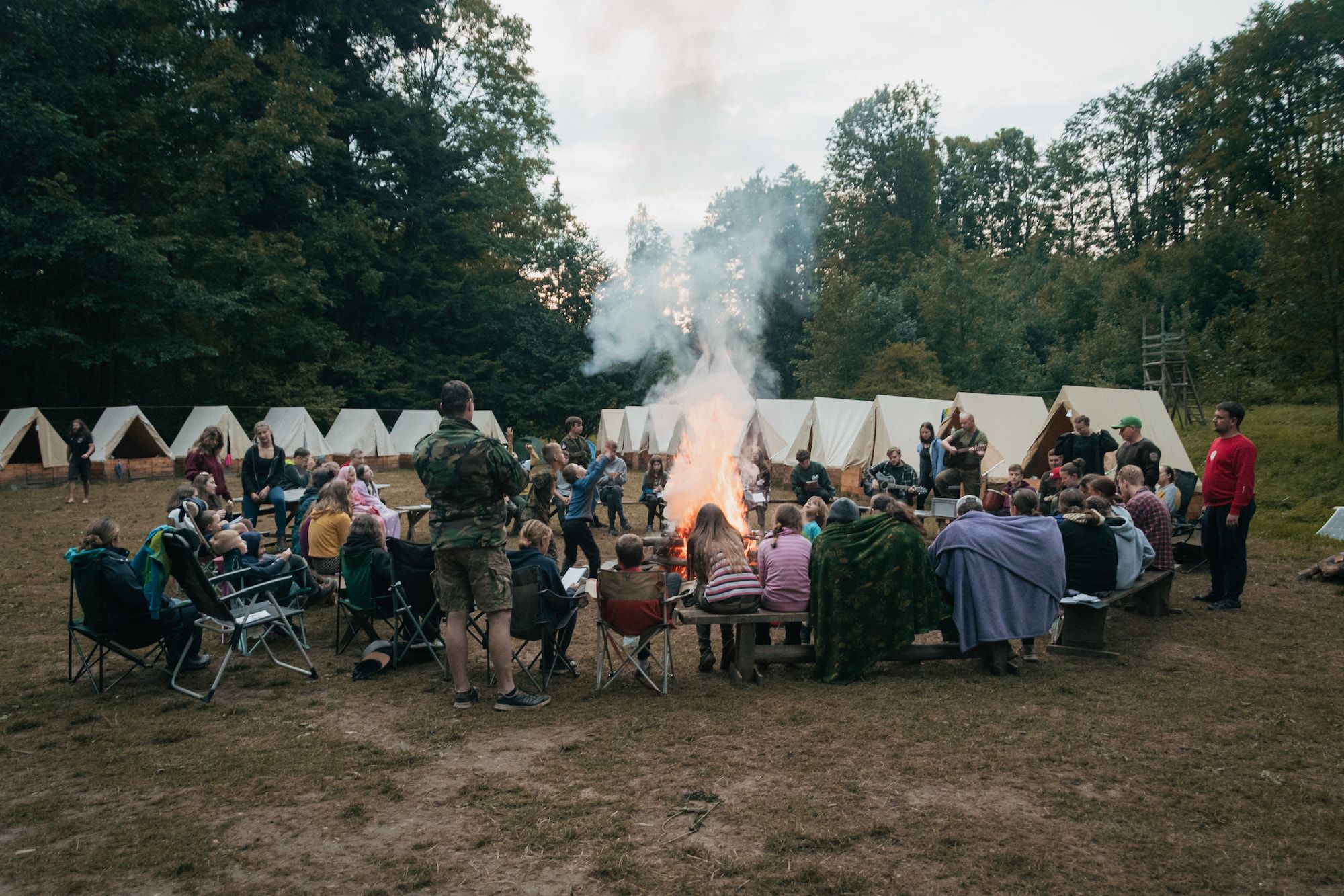 Skupina dětí sedí v kruhu kolem velkého táboráku ve skautském táboře, v pozadí je vidět několik stanů obklopených stromy.