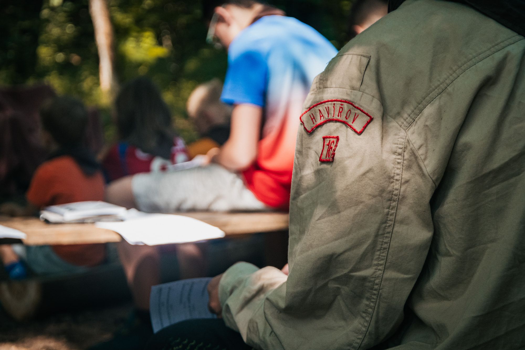 Skaut ve skautské khaki košili s nášivkou „Havířov“ sedí venku a drží v ruce papíry, zatímco v rozmazaném pozadí je vidět několik dětí.